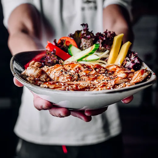 Eine Schale mit gegrilltem Huhn, Gemüse und Salat wird nach vorn gereicht