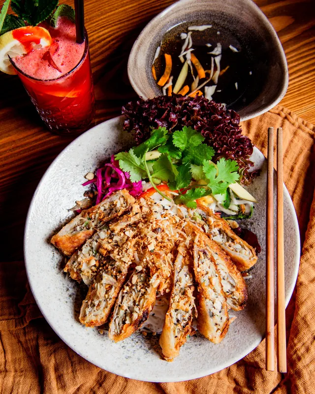 Knusprige Frühlingsrollen in Scheiben auf Reisnudeln mit Koriander, rotem Salat und eingelegtem Rotkohl, daneben Soße und Stäbchen